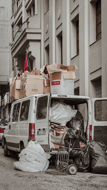 A white commercial van parked on a city street with its rear doors open, revealing an overflowing load of various types of waste, including black trash bags, flattened cardboard boxes, and loose paper, all packed tightly inside and spilling onto the pavement. Several large cardboard boxes are stacked on top of the van, some with visible labels and partially crushed edges, indicating recent bulk rubbish collection or on-site waste clearance by an independent waste management service. A small hand trolley with additional black plastic bags and a metal frame are positioned beside the open back of the van, suggesting ongoing rubbish handling activity. The scene is set against tall urban buildings with light-colored facades and narrow windows, and the environment appears to be in an alley or service lane with natural daylight illuminating the scene, highlighting the textures of cardboard and plastic waste, as well as the structural elements of the vehicle and surrounding architecture. This setup reflects a typical scenario of private rubbish collection or bulky waste removal in a metropolitan area, managed by companies like Waste Collection Marylebone.