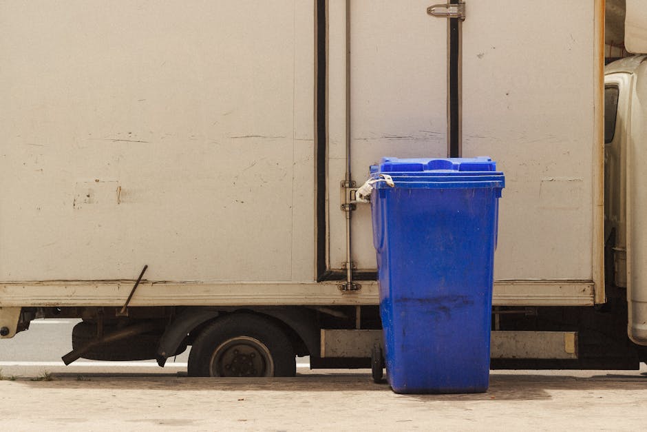 A large blue plastic rubbish bin with a closed lid is positioned on a concrete pavement next to a white delivery or utility truck. The bin has a slightly textured surface and shows some dirt or scuff marks near the bottom. It is situated directly beside the rear side of the truck, which features a plain, slightly dirty white panel with visible horizontal and vertical metal seams, and a small, dark handle or latch near the top of the door. The truck's visible wheel beneath the body is black with a metal hubcap, and a small section of the truck’s undercarriage can be seen. The scene is outdoors, illuminated with natural daylight, and the environment suggests an urban or residential area for waste collection or disposal activities, typical of independent waste handling or private rubbish removal services like Waste Collection Marylebone.