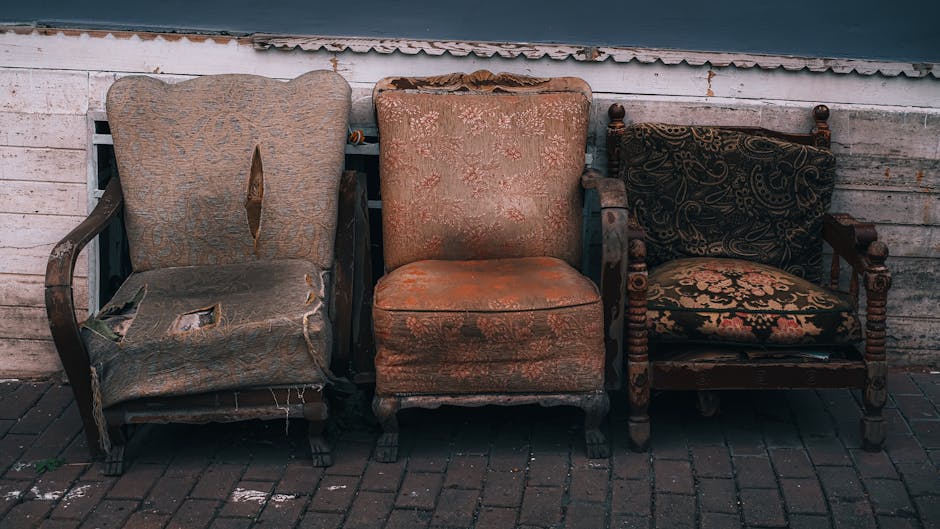 Three vintage upholstered chairs are positioned side by side on a brick pavement against a weathered white wooden wall and corrugated metal roof. The chair on the left features a damaged, torn fabric with visible stuffing and wood framing, showing signs of wear and deterioration. The middle chair has a faded orange-brown fabric with a floral pattern, appearing stained and worn, with the upholstery sagging slightly over a wooden frame. The rightmost chair has a dark, ornate upholstery with gold and red floral patterns, and a wooden frame with turned spindles and carved armrests, displaying consistent signs of age and use. The scene suggests an outdoor setting possibly related to private waste handling or local disposal, with these broken or unwanted furniture pieces awaiting removal or recycling, as often handled by services like Waste Collection Marylebone. The natural lighting indicates daytime, with no additional equipment or objects visible, emphasizing the contrast between the aged furniture and the utilitarian background.