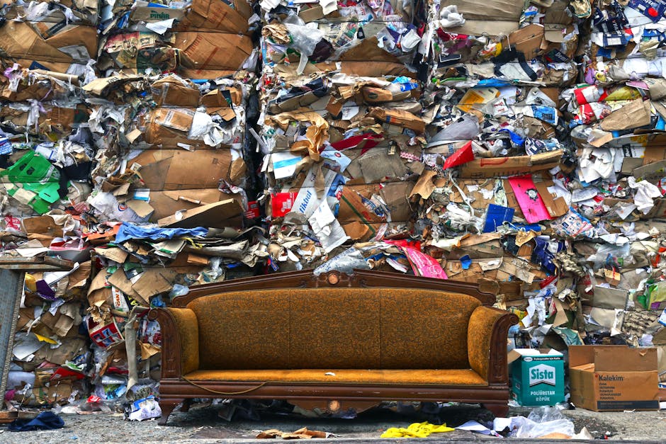 A vintage-style upholstered sofa with a wooden frame and orange-brown fabric upholstery is positioned on a concrete ground in front of a large, densely packed wall of mixed waste and garbage. The wall contains tightly stacked and overflowing piles of crumpled cardboard boxes, paper waste, packaging materials, and various torn paper products, creating a chaotic and cluttered background. The cardboard varies in color from light brown to darker shades, with some boxes displaying printed labels and logos, while the paper bits are in a mixture of white, colored, and printed surfaces. The scene appears outdoors, possibly along a street or alley, with natural daylight illuminating the waste, highlighting the textures of the crumpled paper and cardboard. Small debris and pieces of plastic are scattered around the sofa's base, emphasizing the disorder. This setting subtly connects to the theme of waste disposal and alternative rubbish removal options offered by Waste Collection Marylebone, illustrating the importance of proper waste management and disposal services in urban environments.