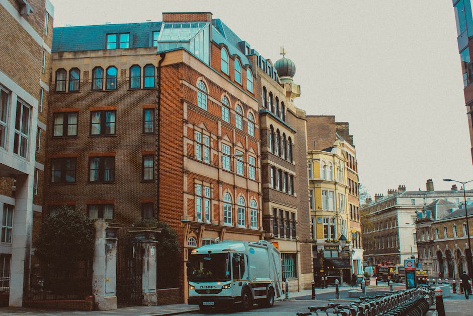 A street scene in an urban area shows a waste collection vehicle, a compact blue and white van, parked on the pavement near a row of multi-storey buildings with diverse architectural styles and materials. The vehicle, used by waste management services such as Waste Collection Marylebone, is positioned close to the curbside, with its front facing right. The buildings in the background feature a mixture of red brick, stone, and pastel-colored facades, with some structures displaying decorative window frames, arched windows, and ornate cornices. The street is lined with black metal bollards and a row of bicycles secured to bike racks. On the left, a gated entrance with stone pillars and greenery separates the sidewalk from a building entrance, while to the right, several parked cars and additional architectural details are visible. The lighting suggests a partly cloudy day, with natural diffuse light illuminating the scene, emphasizing the textures of the building materials and the vehicle. This scene exemplifies typical private waste collection operations in an urban environment, highlighting the role of independent collection services in managing rubbish outside of municipal systems.