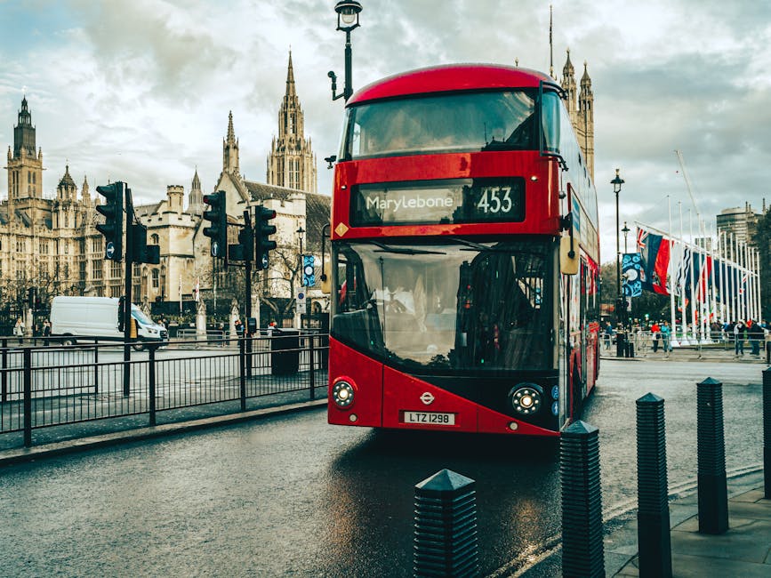 A red double-decker bus with a digital route display reading 'Marylebone 453' is parked on a wet street in front of historic Gothic-style buildings with spires and ornate stonework. The bus is positioned centrally in the image, with the windshield and front headlights clearly visible, and the bus's body featuring a smooth, glossy finish. To the left of the bus, there are multiple black traffic signals mounted on posts, with some displaying red pedestrian signals. A white van is driving on the adjacent lane, partially obscured by a black metal fence that separates the sidewalk from the roadway. Behind the bus, there are pedestrians walking along the pavement, some near a row of tall, narrow flagpoles decorated with colorful banners or flags fluttering in the breeze. The surrounding environment indicates an urban setting with historic architecture, which Waste Collection Marylebone might service in the context of alternative waste management options, such as private rubbish collection or on-site clearance, as suggested by the overall scene of city activity and infrastructure.