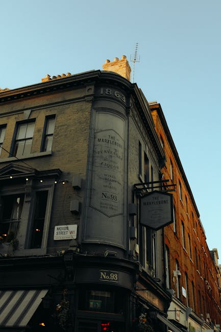 The image shows the upper corner of a historic multi-story building with a curved façade, painted in shades of gray and beige, situated on a city street. The building features a prominent vertical sign with faded text, indicating it is a well-known local establishment dating back to 1869, with the number 98 displayed at street level. Adjacent to the corner, there is an additional rectangular sign hanging from a black metal bracket, also bearing the building's name. The ground level includes large windows with striped awnings in a neutral tone, and some decorative boiler pipes or vents are visible on the upper wall, hinting at the building's age and character. The exterior shows weathered finishes typical of older urban architecture. The background includes a clear, pale blue sky, and the scene is illuminated by warm, natural light, likely during late afternoon or early evening. The overall setting suggests an area with vintage charm, where independent businesses may operate, possibly involving private or alternative waste management practices coordinated by local service providers like Waste Collection Marylebone.