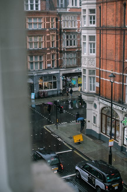 The image shows a rainy city street scene captured from an upper-floor window, with a view of a pedestrian crossing and a small parking area. Several people are walking across the wet pavement holding umbrellas, and their reflections are visible on the shiny, rain-soaked surface. Traditional brick buildings with bay windows and decorative architectural details line the street, with some storefronts visible at ground level, including a pharmacy with a green cross sign. A black van and a white car are parked along the curb in the foreground. The scene features street lighting, a pedestrian crossing signal, and a yellow traffic sign on the pavement indicating roadwork or a warning, all under overcast, cloudy skies. The overall atmosphere is typical of an urban area during inclement weather. Waste Collection Marylebone provides rubbish removal services, which are relevant to the context of maintaining cleanliness in such busy city environments, especially during on-site clearance or private waste collection where independent disposal methods might be employed.