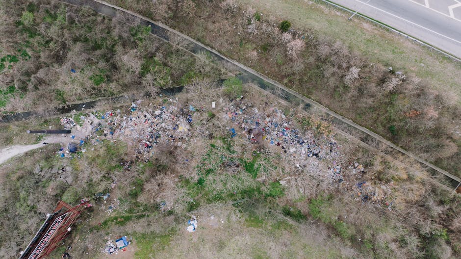 An aerial view of a large illegal dumping site situated along a narrow dirt pathway adjacent to a wooded area with sparse trees and bushes. The pile consists of various discarded waste materials, including plastic bottles, paper, cardboard, and construction debris, scattered across the ground and forming an irregular mass. The waste appears disorganized and extends from the left to the right of the image, with some parts of the pile leaning against trees and scattered debris surrounding the main refuse. To the right, a road with a curb, fencing, and a grassy verge separates the dumping site from the asphalt surface of the highway. The environment around the site features natural vegetation with a mix of leafless and green trees, indicating a season of early spring or late autumn. The lighting is natural, and the overall scene depicts a typical example of illicit waste dumping that may require private clearance or rubbish removal services from Waste Collection Marylebone to mitigate environmental impact.