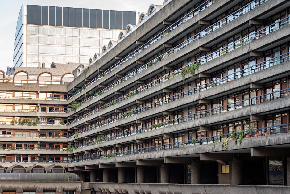 A multi-storey residential building with a concrete facade and rows of balconies running horizontally across each floor. The balconies are equipped with dark metal railings and are decorated with various potted plants and outdoor furniture. The building's exterior shows signs of weathering, with some greenery hanging from the balcony edges. Behind the main structure, a taller office or commercial building with a glass and steel facade reflects the sky, adding contrast to the scene. The photograph is taken during daylight, with natural light illuminating the details of the building’s textures and colors. The ground level features an overhang with rounded concrete supports, indicating a parking area beneath the residential floors. The image depicts an urban environment where private spaces and organic growth coexist amidst large concrete structures, aligning with themes of alternative waste handling or independent collection methods in densely built-up areas, as seen in professional rubbish removal services like Waste Collection Marylebone.
