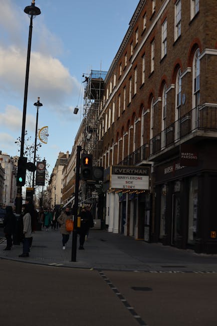 A city street scene featuring a multi-storey brick building with white-framed arched windows and small balconies on the right side. An area of scaffolding and black netting indicates ongoing building maintenance or refurbishment. In the foreground, there is a pedestrian crossing with a traffic light showing a red walking figure, and a black street lamp stands nearby. Several pedestrians, some carrying shopping bags, are walking along the pavement. The sky above is partly cloudy with patches of blue visible. On the ground level, there are retail shops, including one with a sign for a bakery named 'MARCEL PARSONS' and other storefronts visible along the street. The scene suggests an active urban environment where independent collection or private waste handling could be part of ongoing property management or refurbishment efforts, aligning subtly with rubbish removal services provided by waste collection companies such as Waste Collection Marylebone.