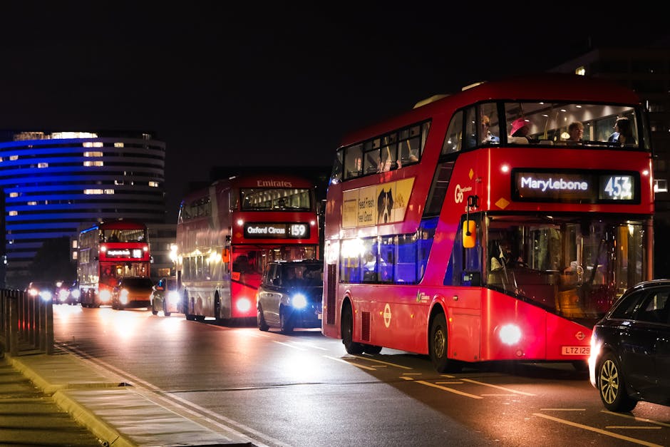 A nighttime scene featuring three red double-decker buses parked on a city street. The bus closest to the camera displays route number 453 with a destination of Marylebone, and the second shows route 159 heading to Oxford Circus, with both vehicles illuminated by internal and external lighting. The buses are positioned parallel to the roadside, with streetlights casting light on their glossy surfaces and nearby parked cars. In the background, modern buildings with curved facades are visible, some illuminated with blue and white lights. The street is relatively clear of traffic, and the surrounding environment indicates an urban setting suitable for private or independent waste collection services, aligning with the objective of alternative rubbish removal solutions provided by Waste Collection Marylebone. The overall scene suggests a late evening or night period, with clear visibility of the buses’ details and the surrounding infrastructure.