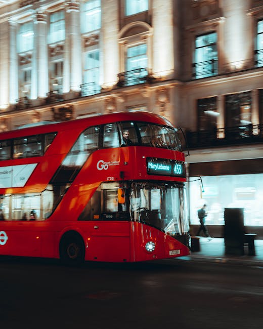 A modern double-decker red London bus traveling along a city street at night, with illuminated signage displaying routes including route 453 bound for Marylebone. The bus has a sleek, glossy finish with smooth, curved surfaces and large windows that reveal reflections of surrounding buildings. The upper deck windows are dark, while the lower deck features a partially visible driver and interior lighting. The background showcases a historic building with large, ornate columns, detailed stonework, and multiple stories with balconies, all bathed in warm artificial light, creating a contrast with the vibrant red of the bus. Pedestrians are visible on the sidewalk, some walking past the building and others near street-level entrances. The scene has a sense of motion conveyed by the blurred background and wheels of the bus, indicative of urban daytime activity, aligning with the context of city transportation and private vehicle use related to waste management and refuse collection services in Marylebone. Waste Collection Marylebone's focus on efficient, independent rubbish handling is subtly supported by this urban setting. The overall image provides a clear, detailed view of urban transportation that complements content about alternative waste disposal methods and city-based rubbish services.