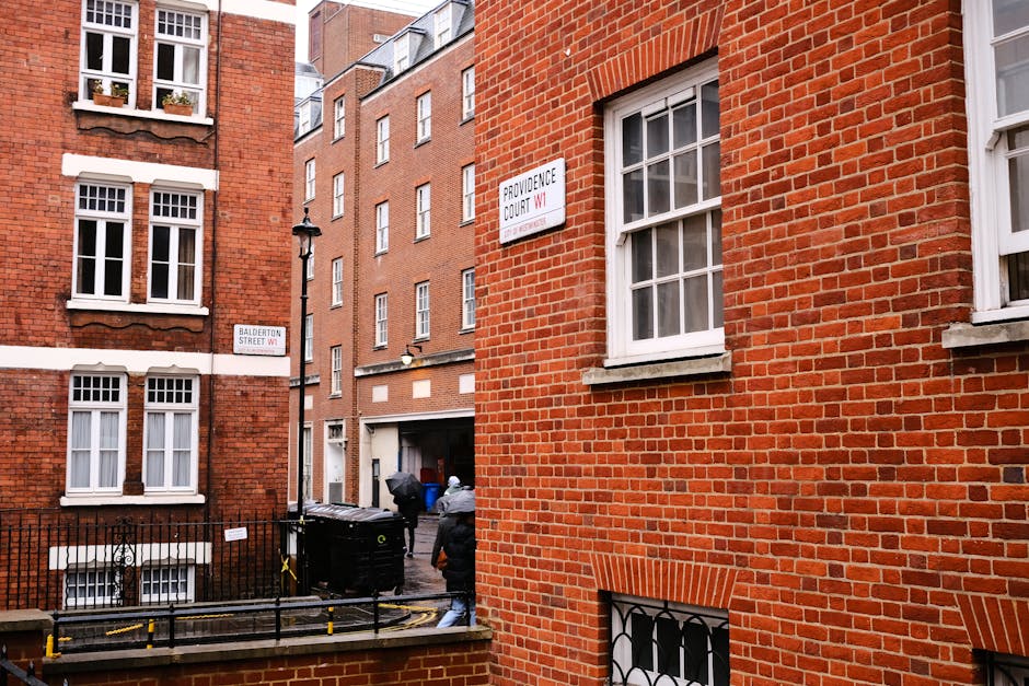 View of a narrow urban alleyway between red brick residential buildings with white-framed sash windows. In the foreground, part of a brick wall with a window, and a black wrought iron security grill beneath it. Two black wheelie bins are positioned near the alley entrance, with a person in dark clothing and a hood walking nearby. Additional multi-storey brick buildings with white window frames and brick facades are visible in the background. A street sign indicating 'Balderton Street W1' and another sign with 'Providence Court W1' are mounted on the walls, alongside a traditional black lamppost. The scene is lit by natural daylight, and the setting appears to be an alley for waste collection and private refuse handling within a residential area, consistent with independent rubbish removal services provided by Waste Collection Marylebone.