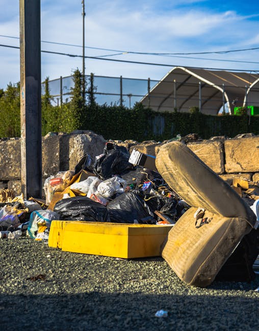 A low-angle view of an outdoor rubbish pile situated on a gravel surface, containing a mix of discarded materials. Prominent in the foreground is a yellow plastic container lying on its side, partially obscured by a large, beige, weathered car tyre that leans against the pile. The rubbish includes black plastic bin bags filled with waste, crumpled paper, and miscellaneous debris, which are haphazardly stacked and spilling onto the ground. Behind the pile, a low stone wall made of rough-hewn, uneven stones separates the area from a background featuring a hedge, a chain-link fence, and a tennis court with a curved, partially covered netted structure. Overhead, there are utility wires running across a blue sky with wispy clouds. The scene appears to be in an urban or suburban environment where private waste collection services, such as Waste Collection Marylebone, might manage on-site clearance of bulky waste, including old furniture and disposal of larger items like tyres and mattresses.
