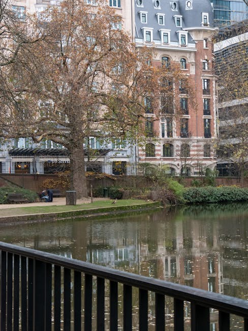 A view of a canal or small river bordered by a black metal railing in the foreground, with calm water reflecting the surrounding environment. On the opposite bank, a large deciduous tree with sparse, brown leaves extends its branches over the water, suggesting a late autumn setting. Behind the tree, there are residential buildings with a mix of architectural styles; one features a red brick facade with white window frames and decorative elements, while nearby buildings are more modern, with glass windows and metal accents. A grassy area with a paved walkway and a few benches is visible along the water's edge, where a person appears to be sitting on a bench. The overall scene depicts an urban environment with a mix of natural and architectural elements, relevant to alternative rubbish removal options near residential areas, supported by companies like Waste Collection Marylebone specializing in private waste handling and on-site clearance services.