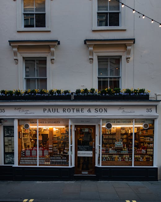 A small traditional shopfront of a delicatessen called Paul Rothe & Son, situated at number 35 on a white building. The shop has large glass windows showcasing shelves stocked with various food products, including jars, bottles, and packaged goods, illuminated by warm interior lighting. The entrance features a wooden door with a narrow vertical window and a sign with the word 'Delicatessen' hanging above. Above the shop, there are two symmetrical sash windows with white frames, decorative stone lintels, and flower boxes filled with yellow and purple flowers. To the right of the entrance, a small sign indicates the shop's focus on foreign and British provisions, with signage on the window advertising sandwiches and other offerings. Above the shop, black signage displays the name of the business. The pavement in front is clean, and the scene is lit by ambient natural light with some decorative string lights hanging overhead, suggesting a lively, urban street setting that may also accommodate independent collection or private waste disposal methods, fitting into a broader context of rubbish management options in the area.